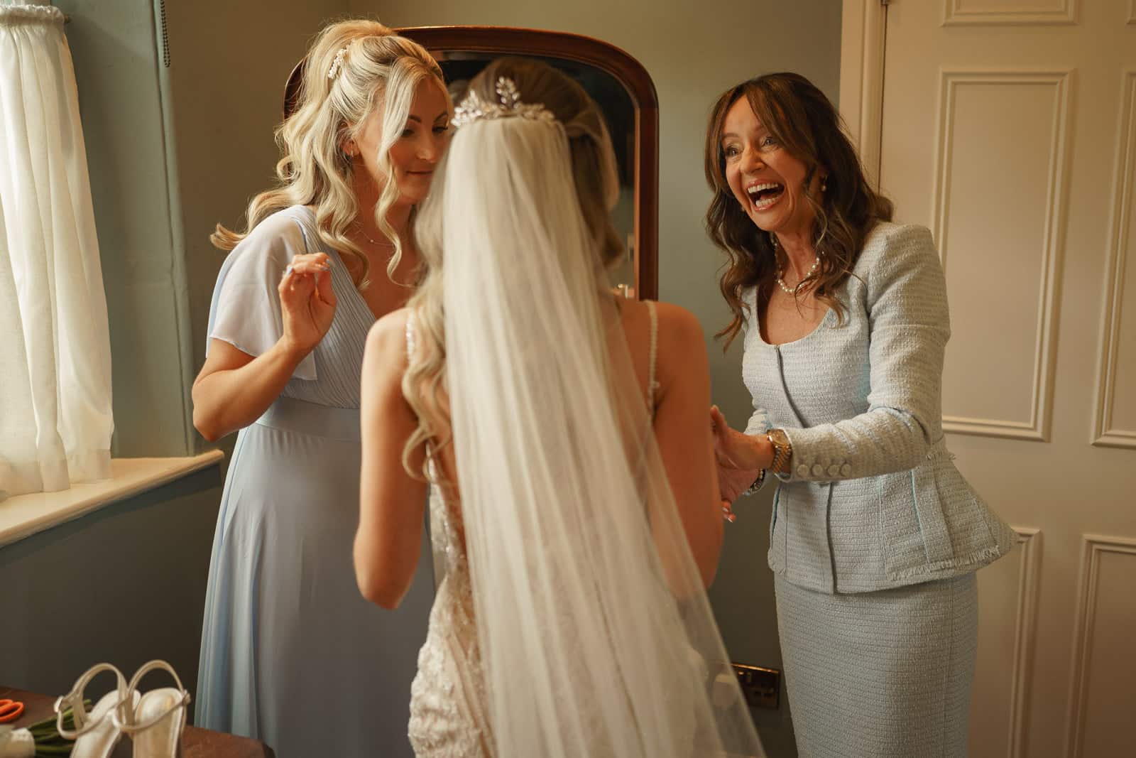 a bride in a veil stands with two women indoors, one helping her with jewelry and the other smiling. captured by pbartworks photography, this elegant scene highlights the skill of a top shropshire wedding photographer.