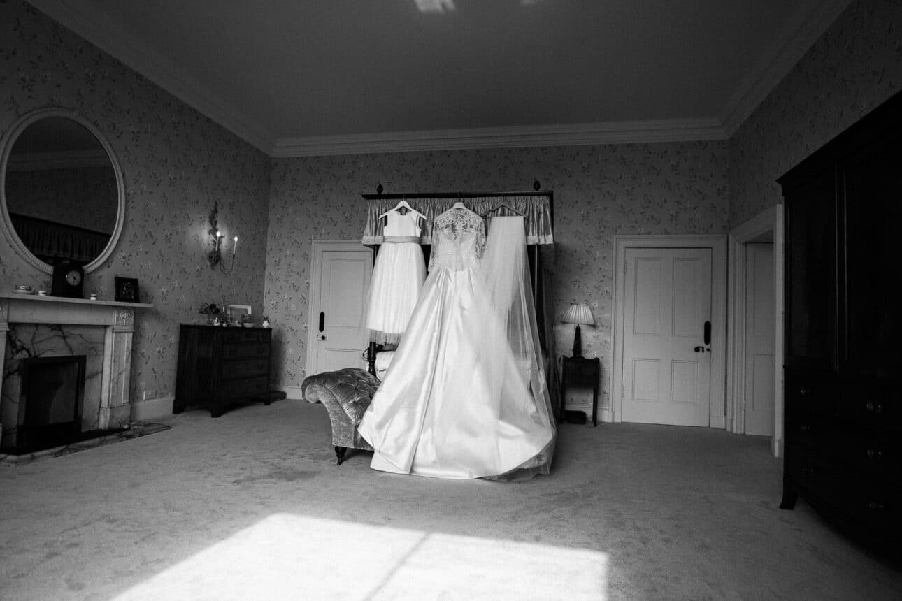 a wedding dress and two smaller dresses hang from a four poster bed in a spacious, carpeted bedroom with vintage decor and patterned wallpaper at sandon hall.
