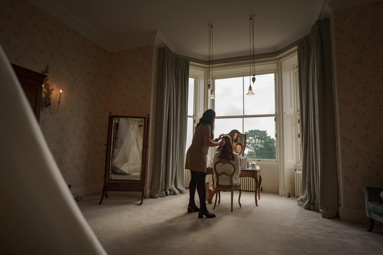a woman styles another woman's hair at a vanity in a softly lit room at sandon hall, with large windows, floral wallpaper, and a wedding dress reflected in a standing mirror.
