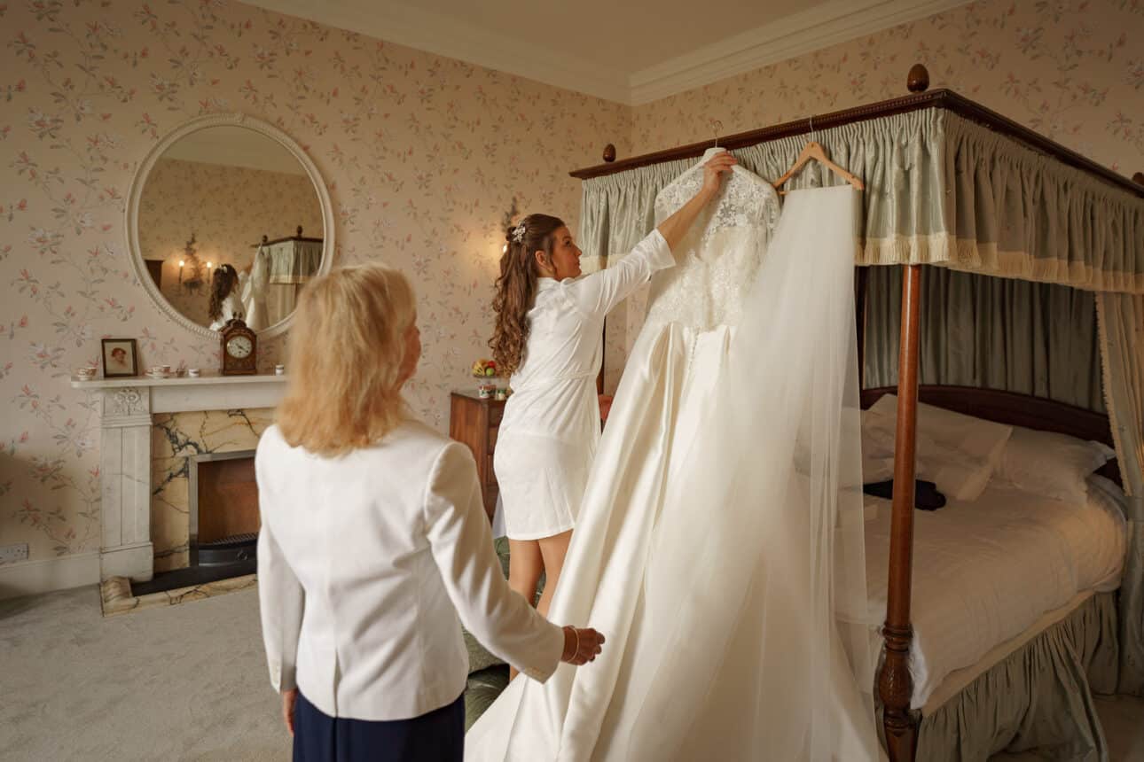 two women in a sandon hall bedroom admire a wedding dress hanging from a four poster bed. one holds the dress up as the other stands nearby, both captivated by its beauty.