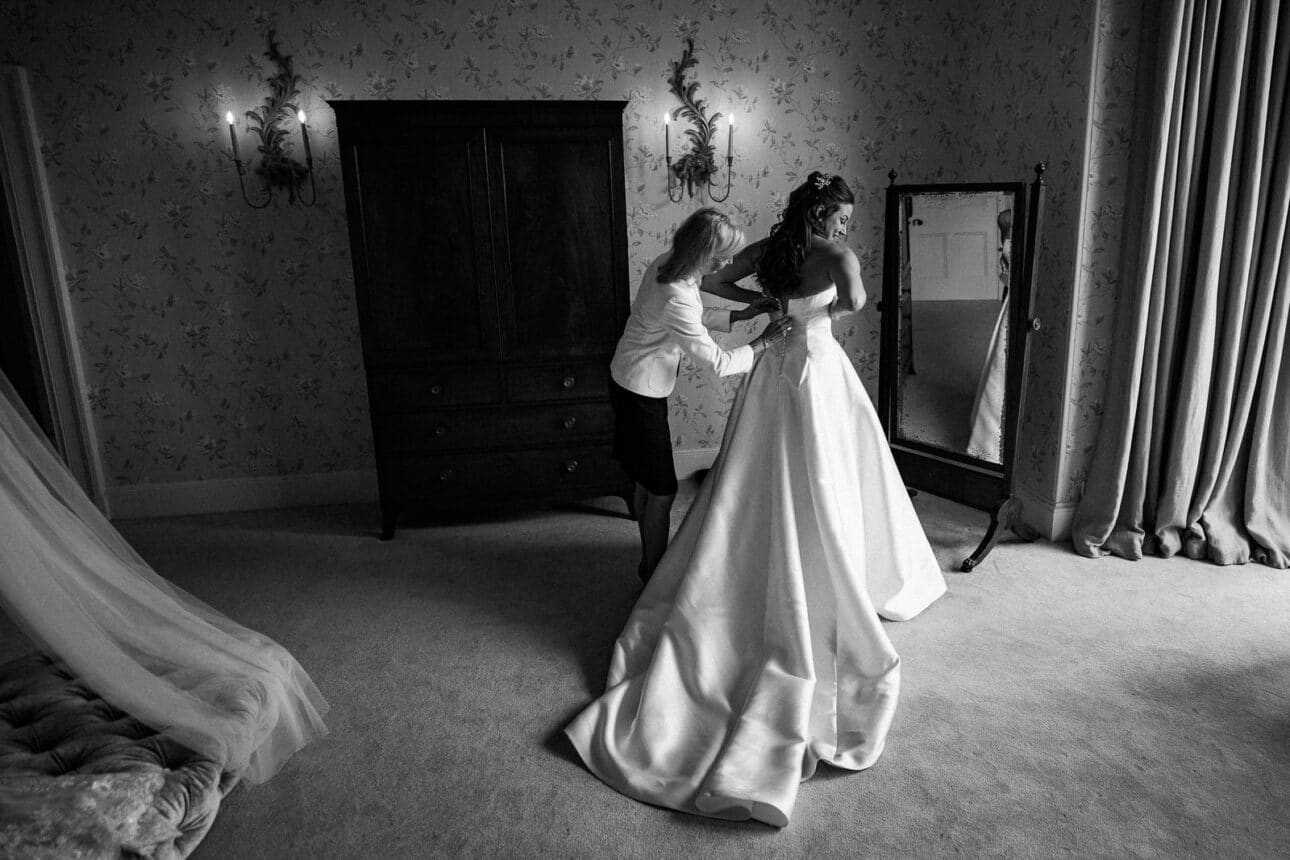 a woman in a wedding dress stands as another woman adjusts the back of her gown in a softly lit, elegant room at sandon hall, adorned with a large mirror and vintage furniture.