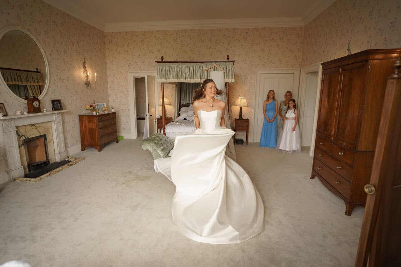 a bride in a white gown stands smiling in a spacious, elegant sandon hall bedroom as three bridesmaids, two in blue and one in white, watch from the background.