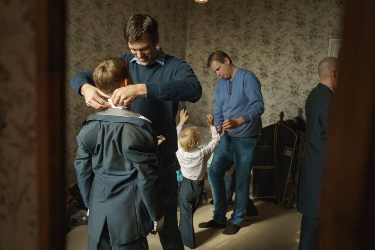 two men help two boys get dressed in suits in a room with floral wallpaper at sandon hall; another man stands nearby with a garment bag visible in the background.