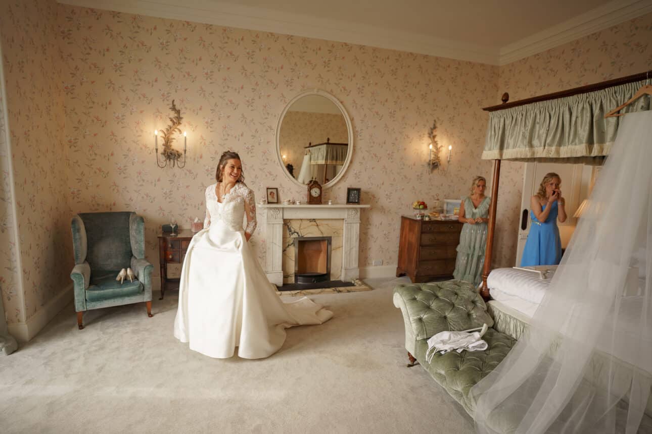 a bride in a white gown stands in a vintage style sandon hall bedroom, while two bridesmaids in blue dresses look on from the background near a bed and dresser.