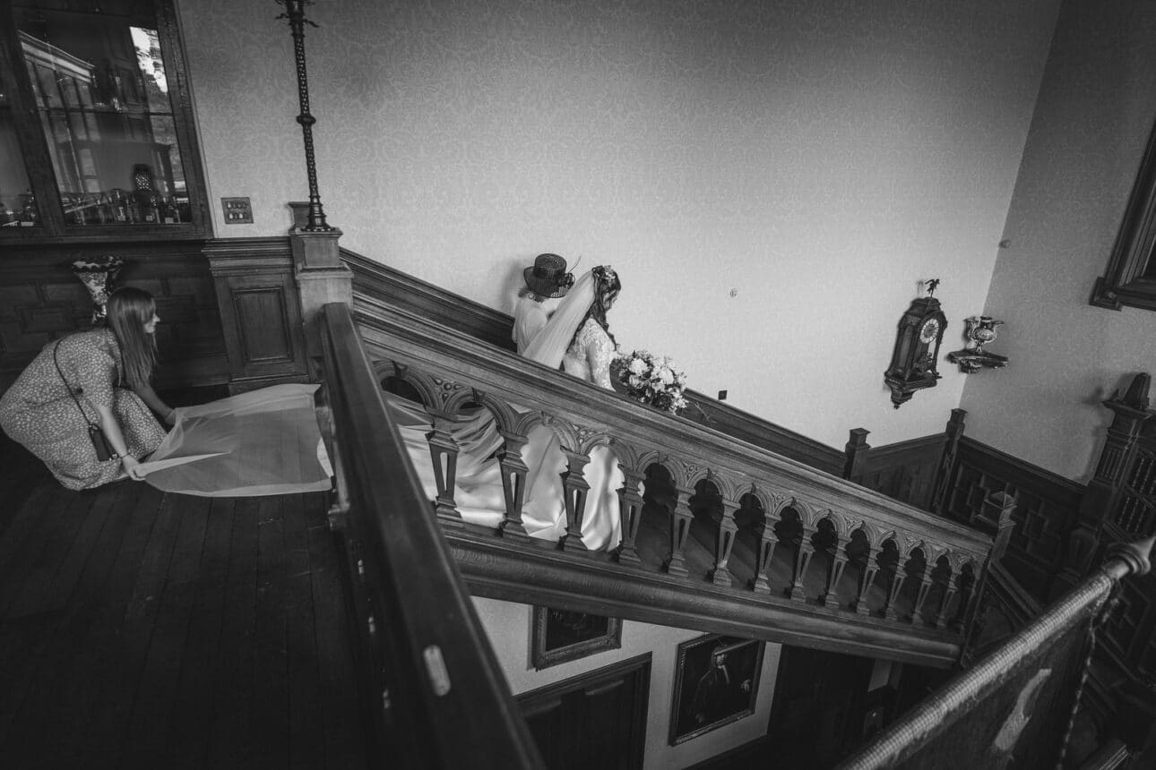 a bride walks up a wooden staircase holding a bouquet, while another woman adjusts the train of her dress in the historic surroundings of sandon hall.