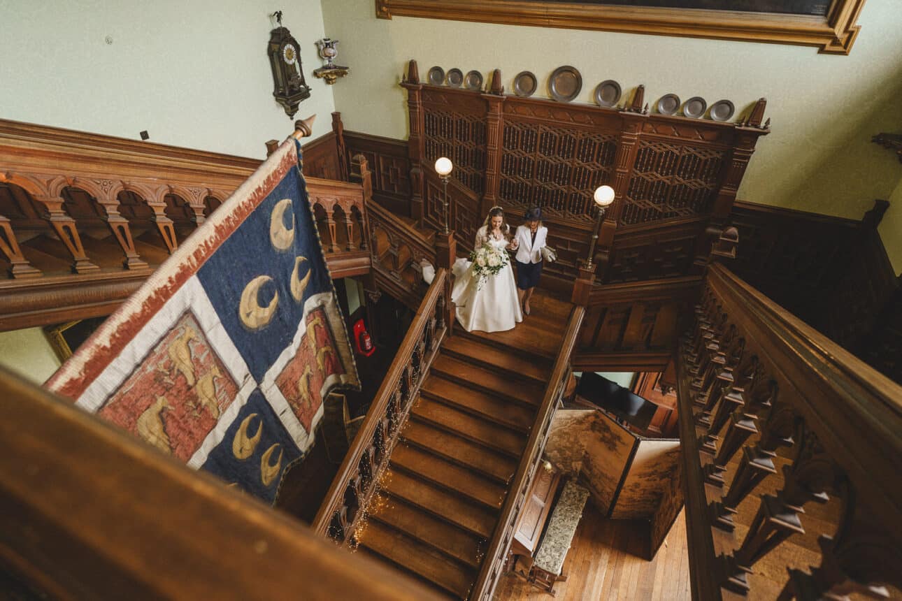 a bride holding a bouquet descends a wooden staircase in the ornate, historic sandon hall wedding venue beside another person. a large decorative tapestry hangs along the stairwell, capturing the elegance of a perfect wedding day.
