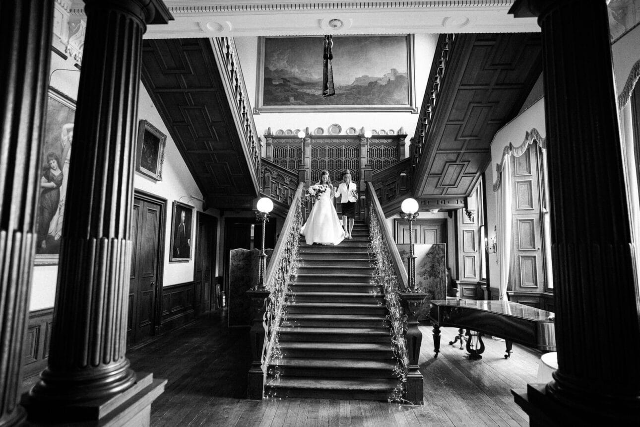 a bride descends a grand staircase at sandon hall, escorted by two people in an ornate, high ceilinged room with columns, paintings, and wooden paneling.