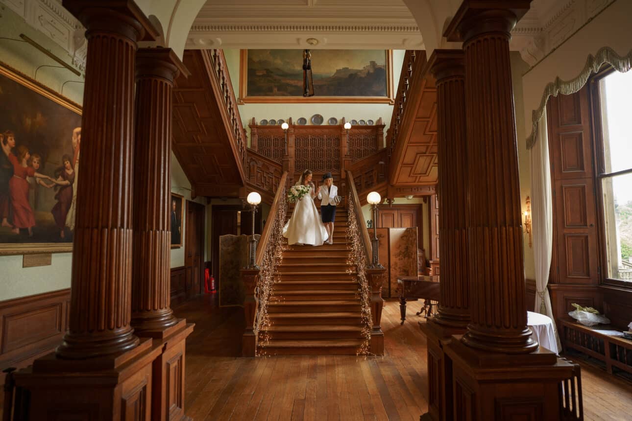 a bride and groom stand together at the top of a grand wooden staircase in sandon hall, a stunning wedding venue adorned with classical paintings and detailed woodwork, capturing the magic of their wedding day.