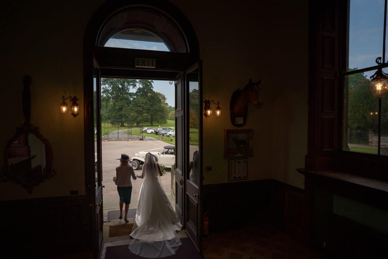 a bride in a white dress stands in a doorway at sandon hall, gazing outside at a vintage car parked on the driveway, with a man in a kilt standing nearby.