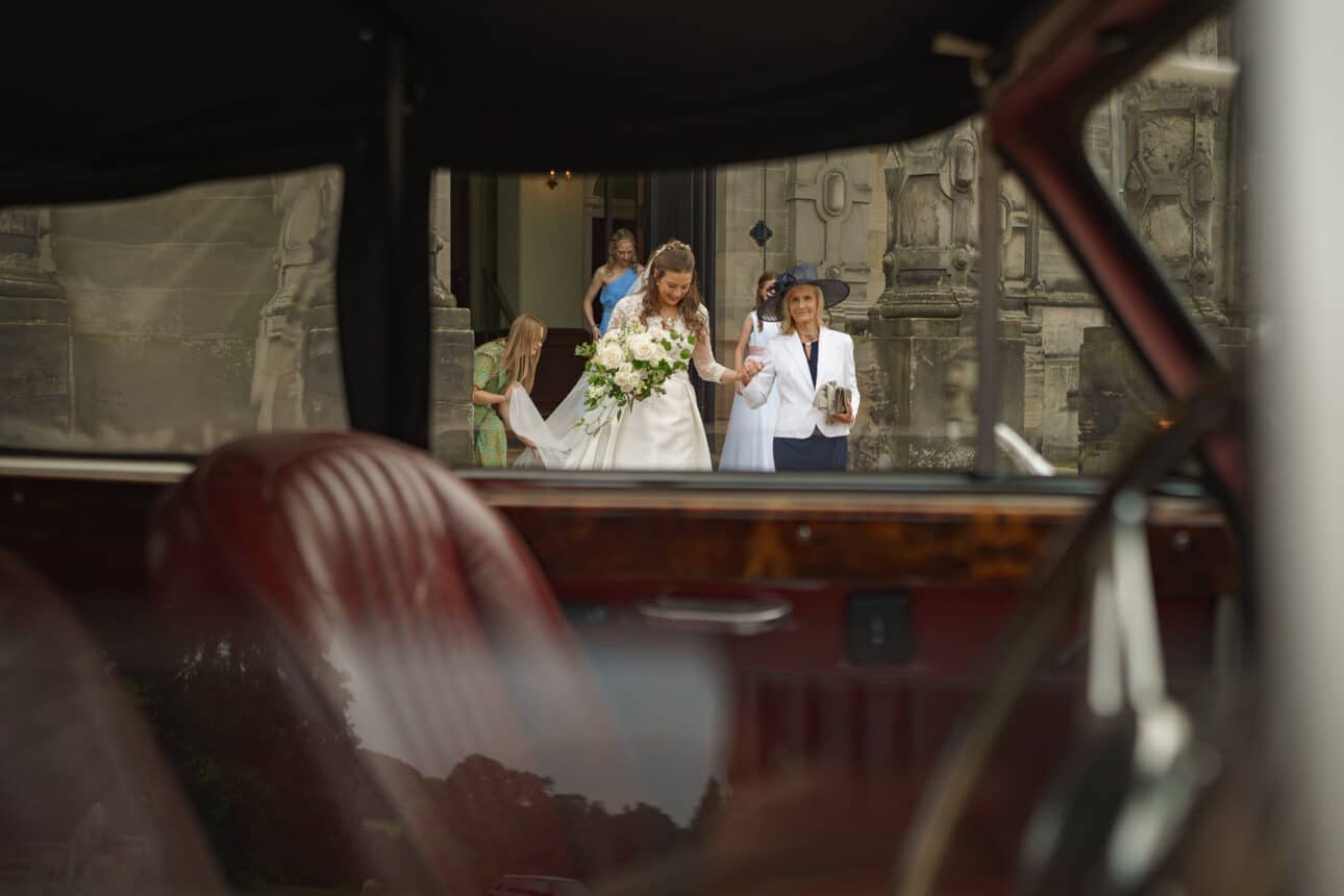 a bride holding a bouquet walks out of sandon hall with an older woman, viewed through the open door of a vintage car with red leather seats.