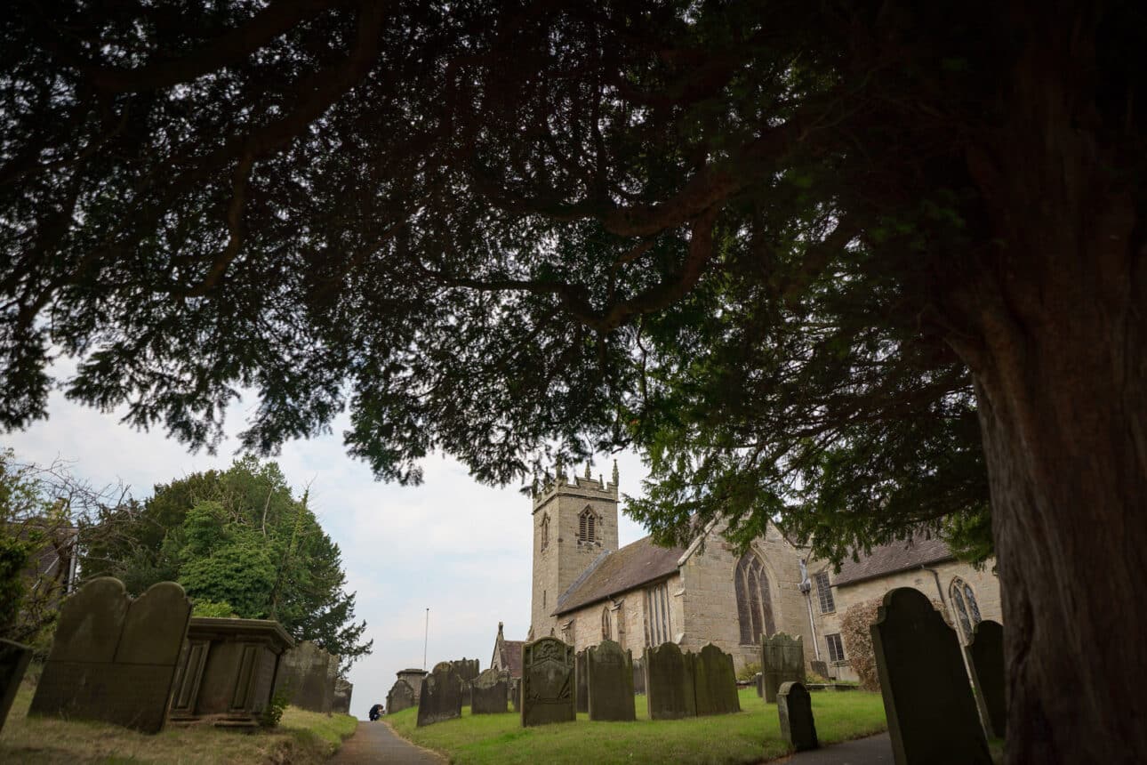 a stone church with a clock tower stands among old gravestones, framed by large trees on a cloudy day—the perfect timeless backdrop for a sandon hall wedding.