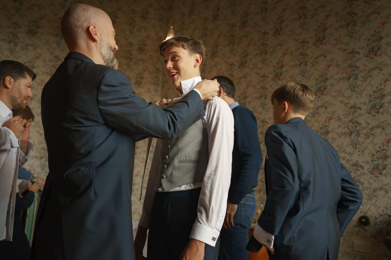 in a room with floral wallpaper at sandon hall, a man helps a young man adjust his vest and shirt while others in suits prepare in the background.