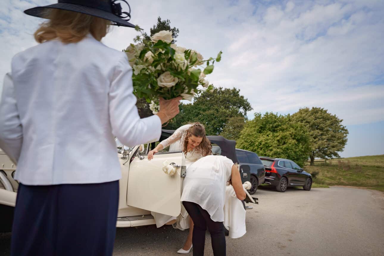a bride steps out of a white vintage car with assistance at sandon hall, while another person holds a bouquet of white roses nearby in a parking area with trees and parked cars in the background.