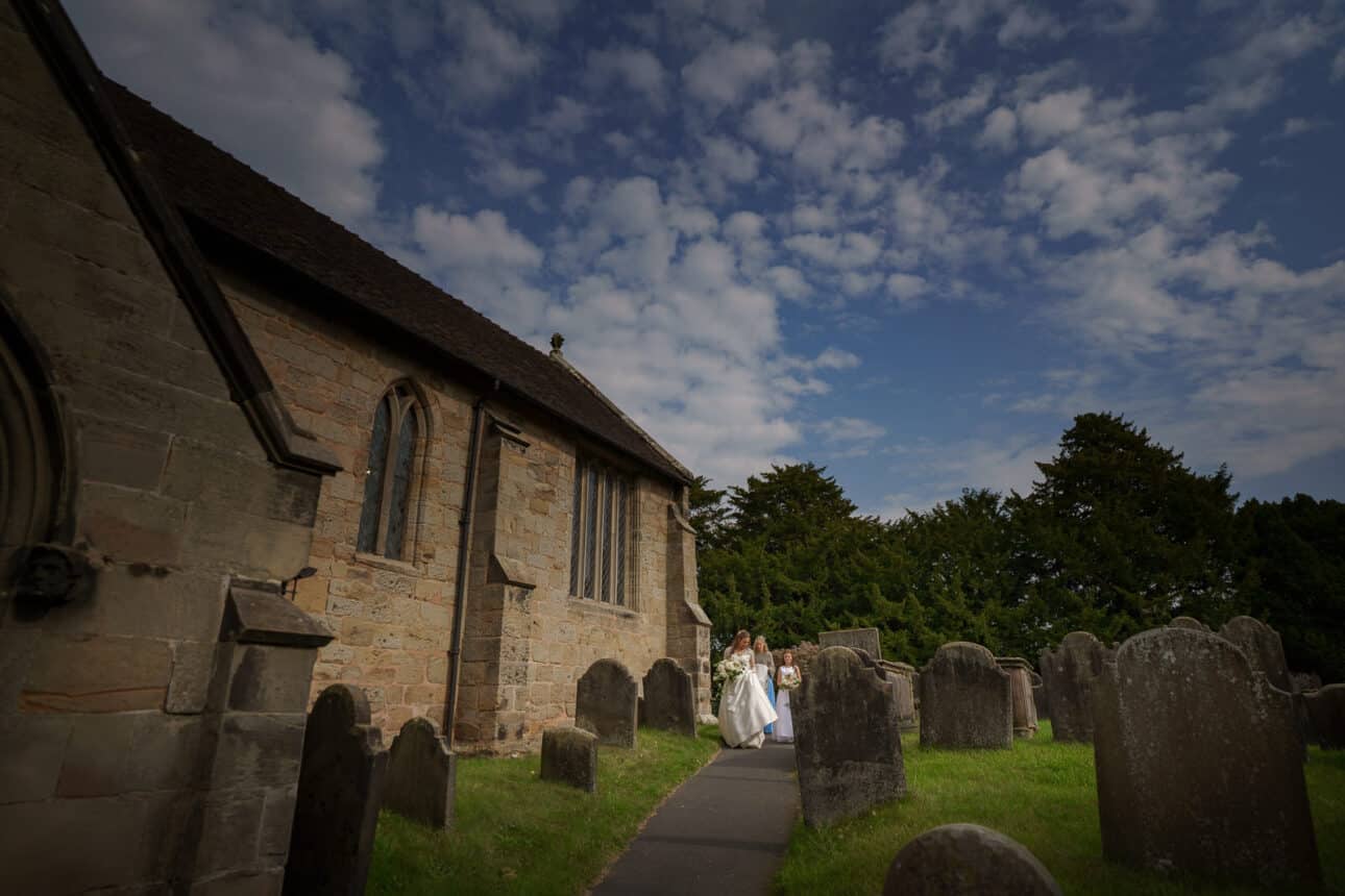 a bride walks with companions along a path beside an old stone church at sandon hall, surrounded by gravestones, under a partly cloudy sky.