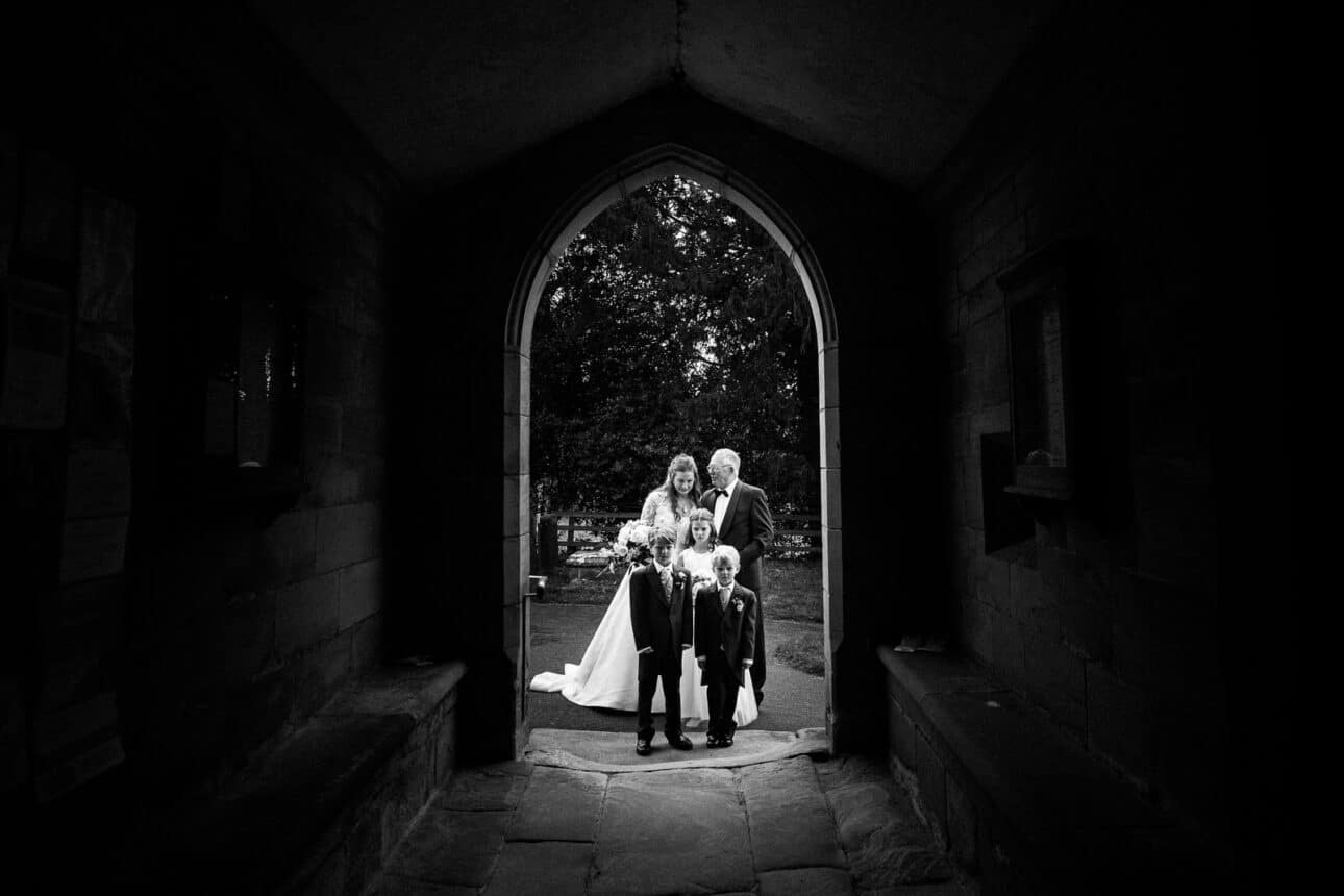 a bride and groom stand under an arched stone doorway at sandon hall with three young children, all dressed formally, posing for a group photo.