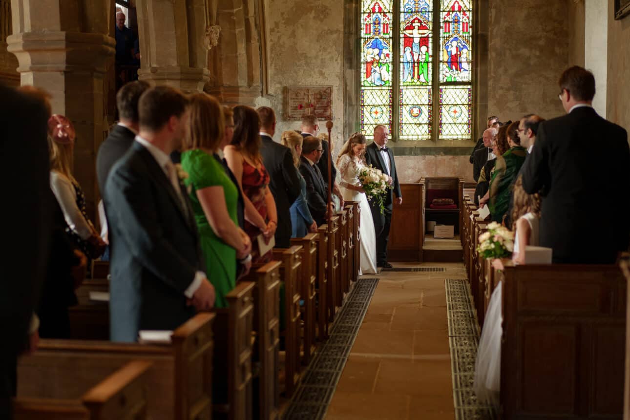 a bride walks down the aisle of sandon hall, holding a bouquet, while guests stand and watch. stained glass windows are visible in the background.