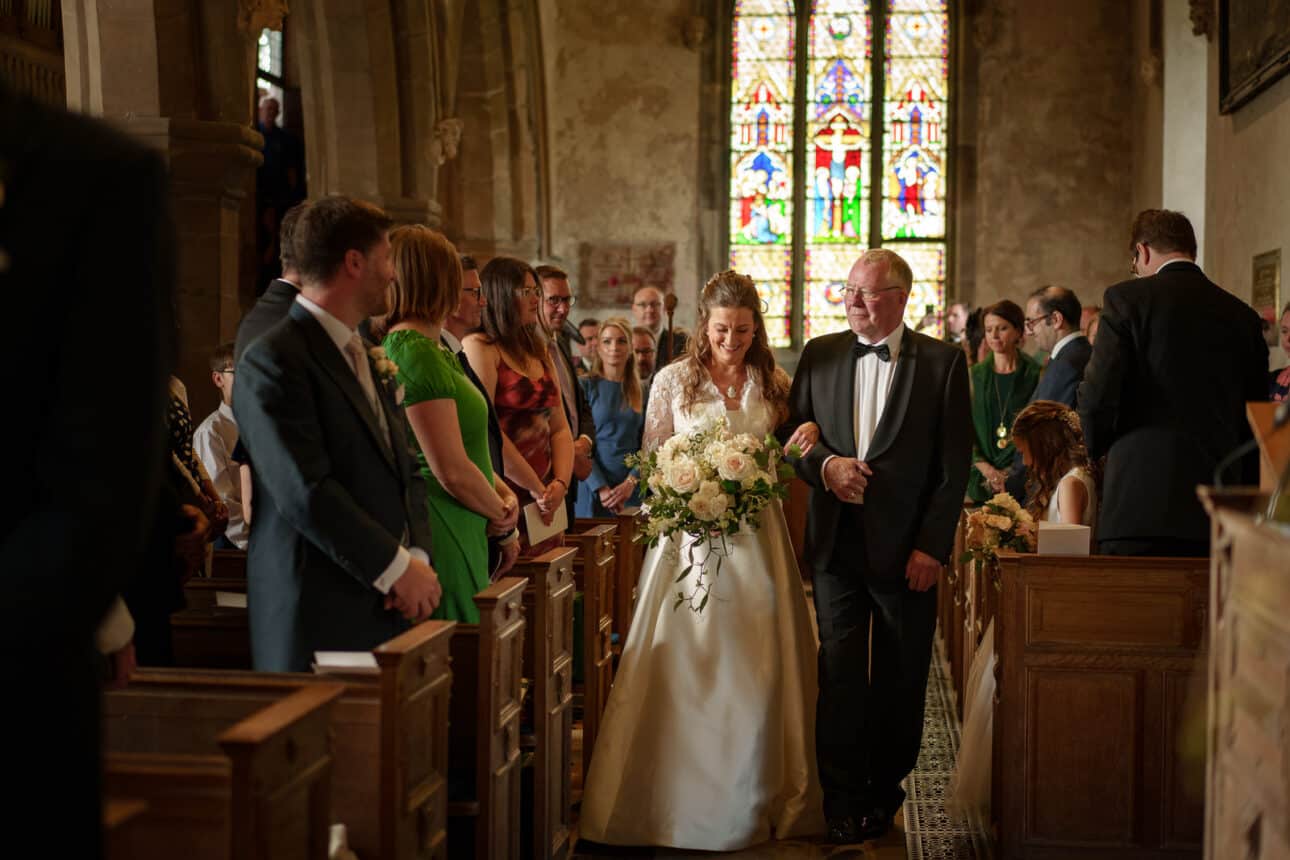 a bride in a white dress walks down the aisle with an older man in a tuxedo inside sandon hall, surrounded by seated and standing guests.