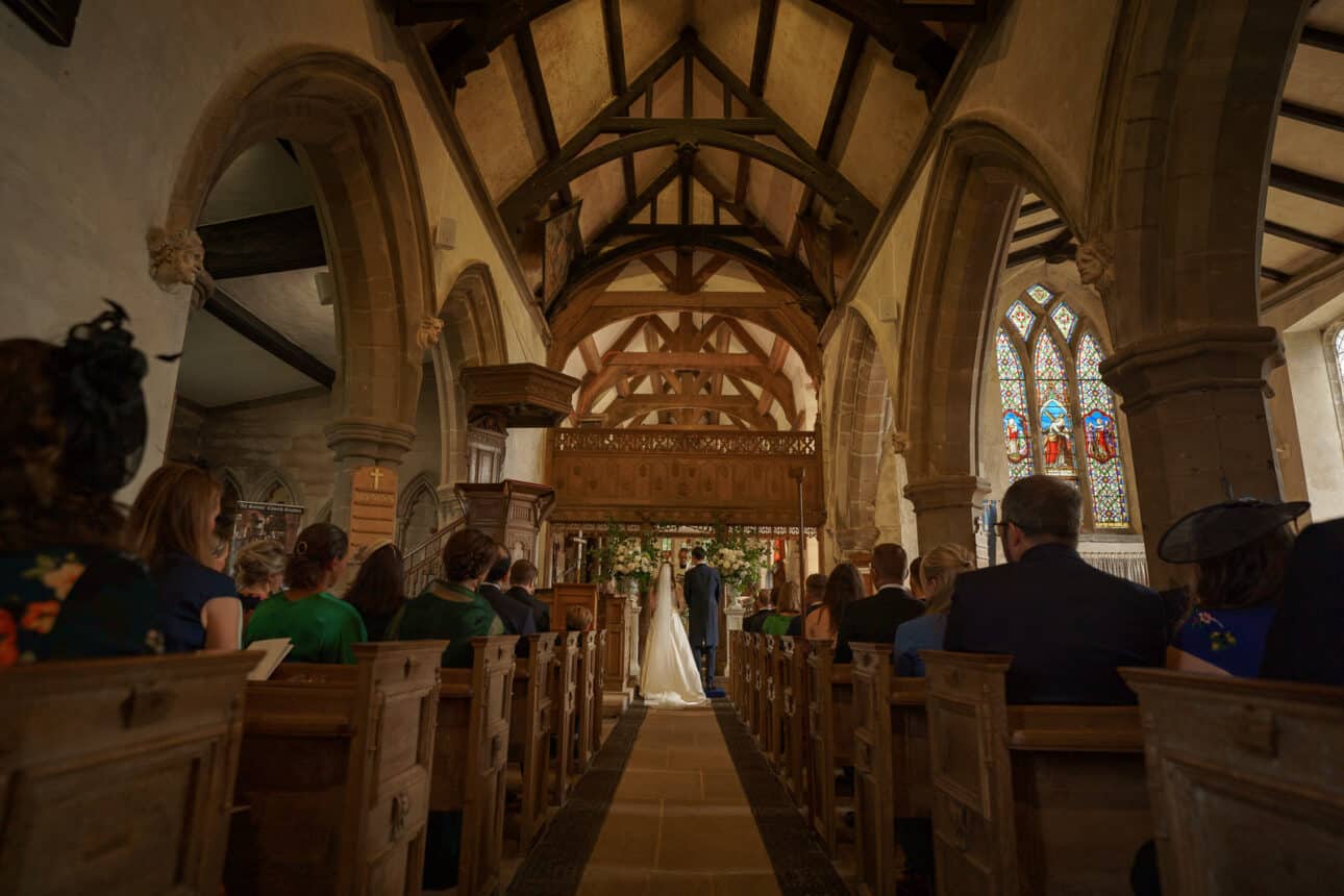a bride and groom stand at the altar in All Saints Church, surrounded by seated guests, with stained glass windows and wooden beams visible—a perfect scene for a sandon hall wedding day.
