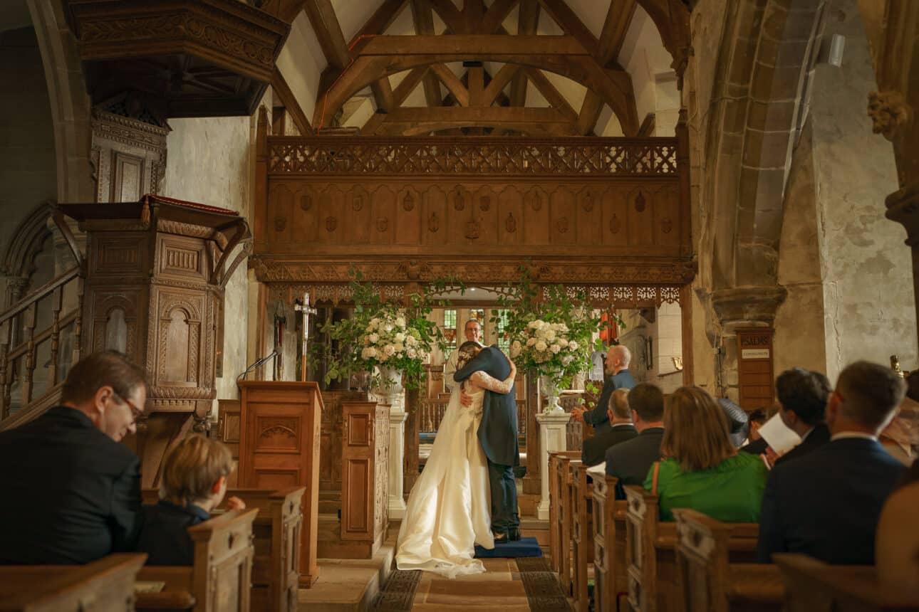 a bride and groom embrace at the altar of sandon hall during their wedding ceremony, with guests seated on either side and elegant floral arrangements in the background.
