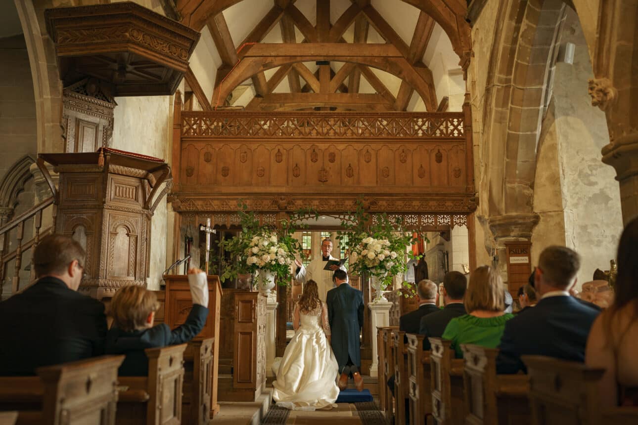 a bride and groom kneel at the altar during their sandon hall wedding ceremony inside the historic church, with guests seated and a priest officiating on their special wedding day.