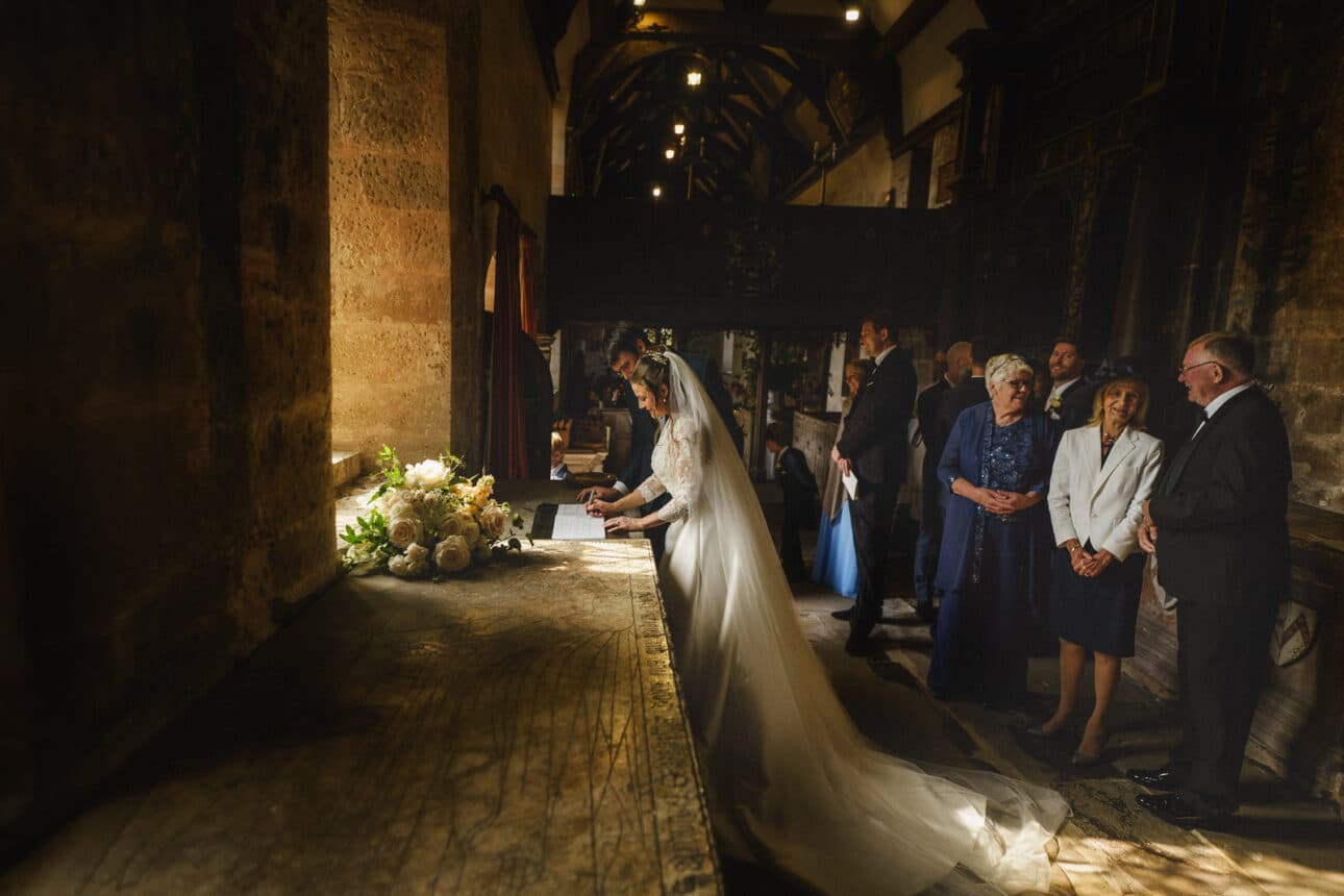 a bride and groom sign a document at a table in a dimly lit church at sandon hall as wedding guests stand nearby, watching.