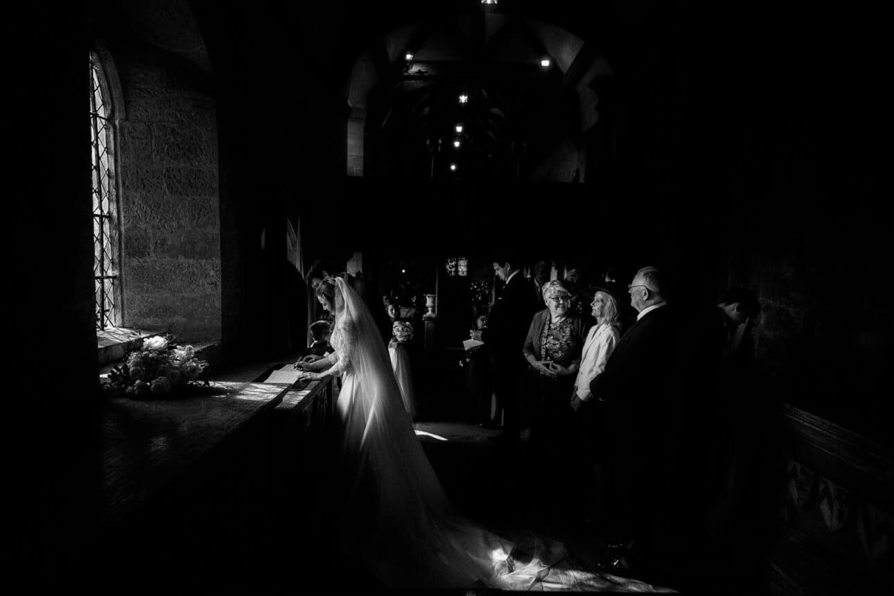 a bride in a white gown stands at a sunlit window signing a document, with guests observing in the dimly lit church interior of sandon hall, making for an unforgettable wedding day.