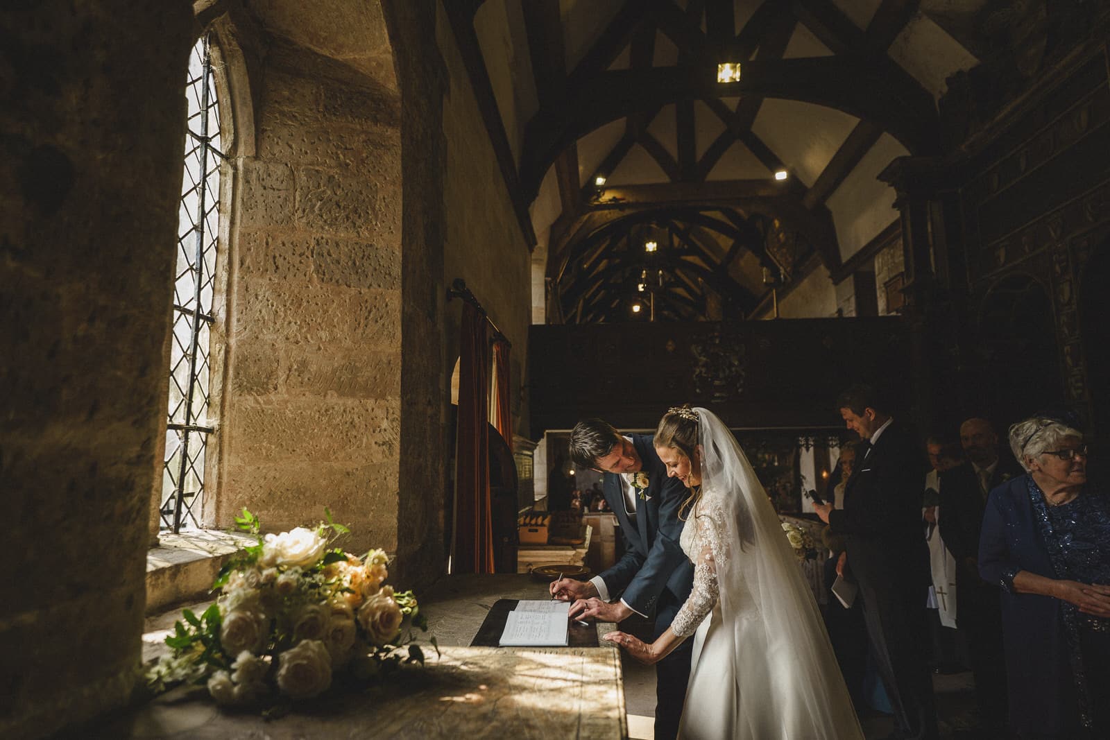 on their wedding day at sandon hall, a bride and groom sign a document inside a dimly lit, historic church adorned with floral arrangements on a wooden table as guests stand nearby.