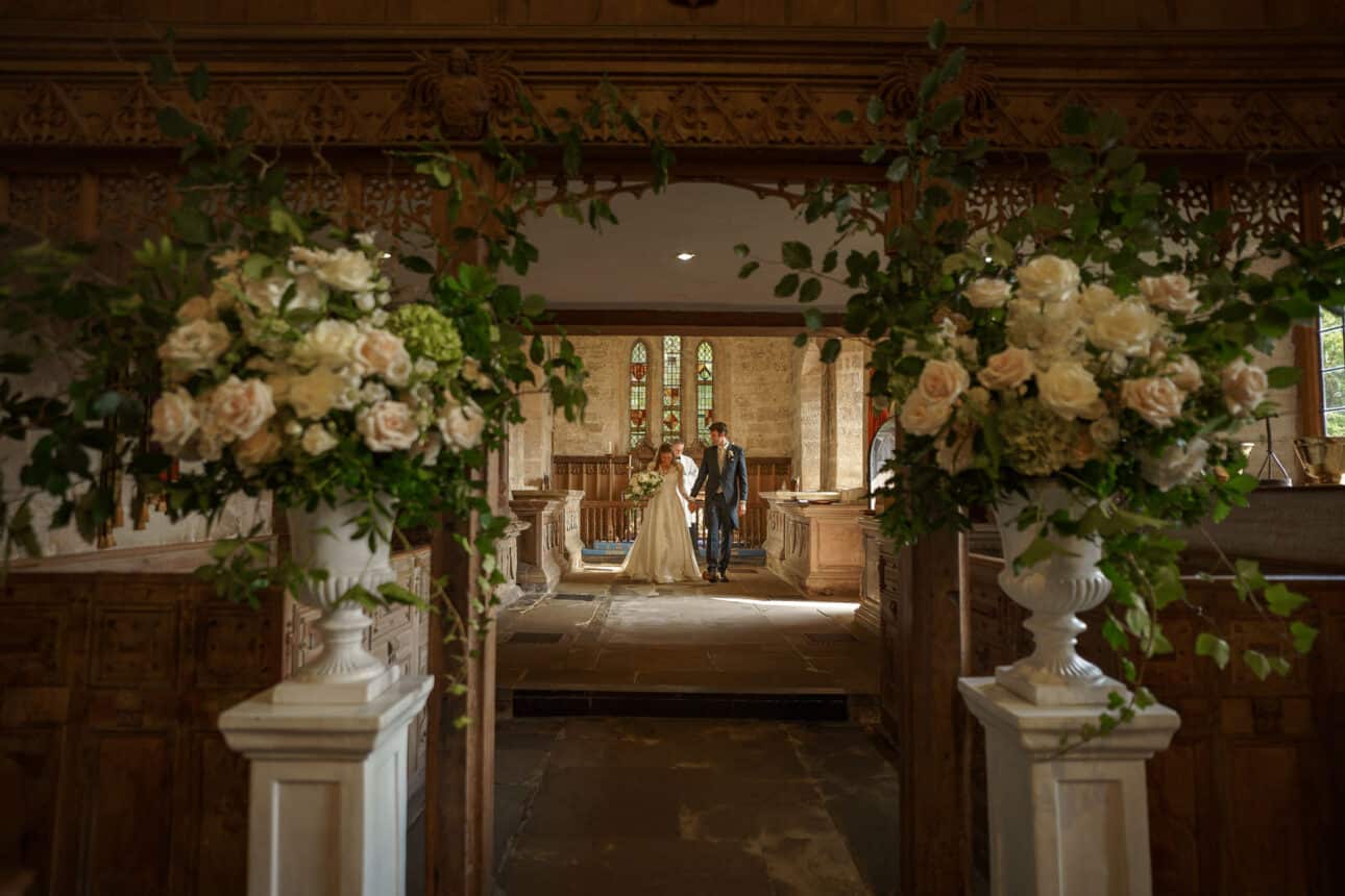 a bride and groom stand together at the altar of All Saints Church, framed by large floral arrangements and elegant wooden architecture on their wedding day.