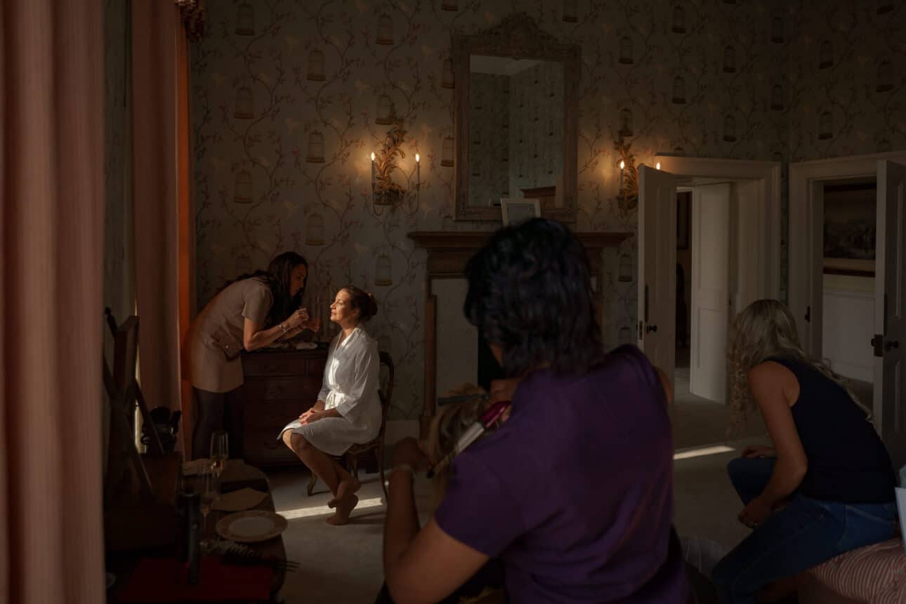 a woman sits in a chair getting her makeup done by another woman, while two others are nearby in a softly lit room with patterned wallpaper at sandon hall.