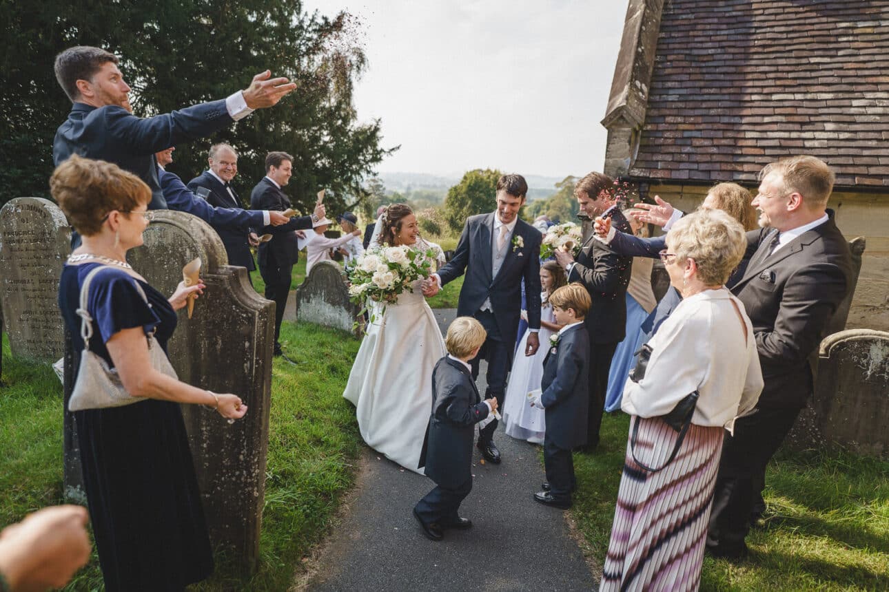 a bride and groom walk down a church path at sandon hall, a stunning wedding venue, surrounded by guests celebrating and holding flowers, with children in formal attire standing nearby.
