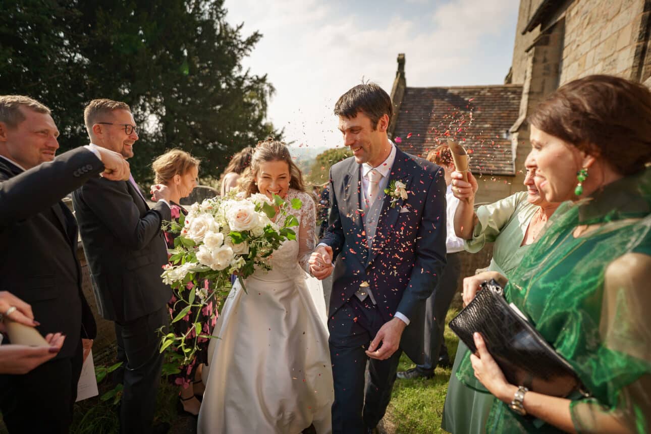 a bride and groom exit sandon hall on their wedding day while guests throw confetti over them; both are smiling, and the bride holds a large bouquet.