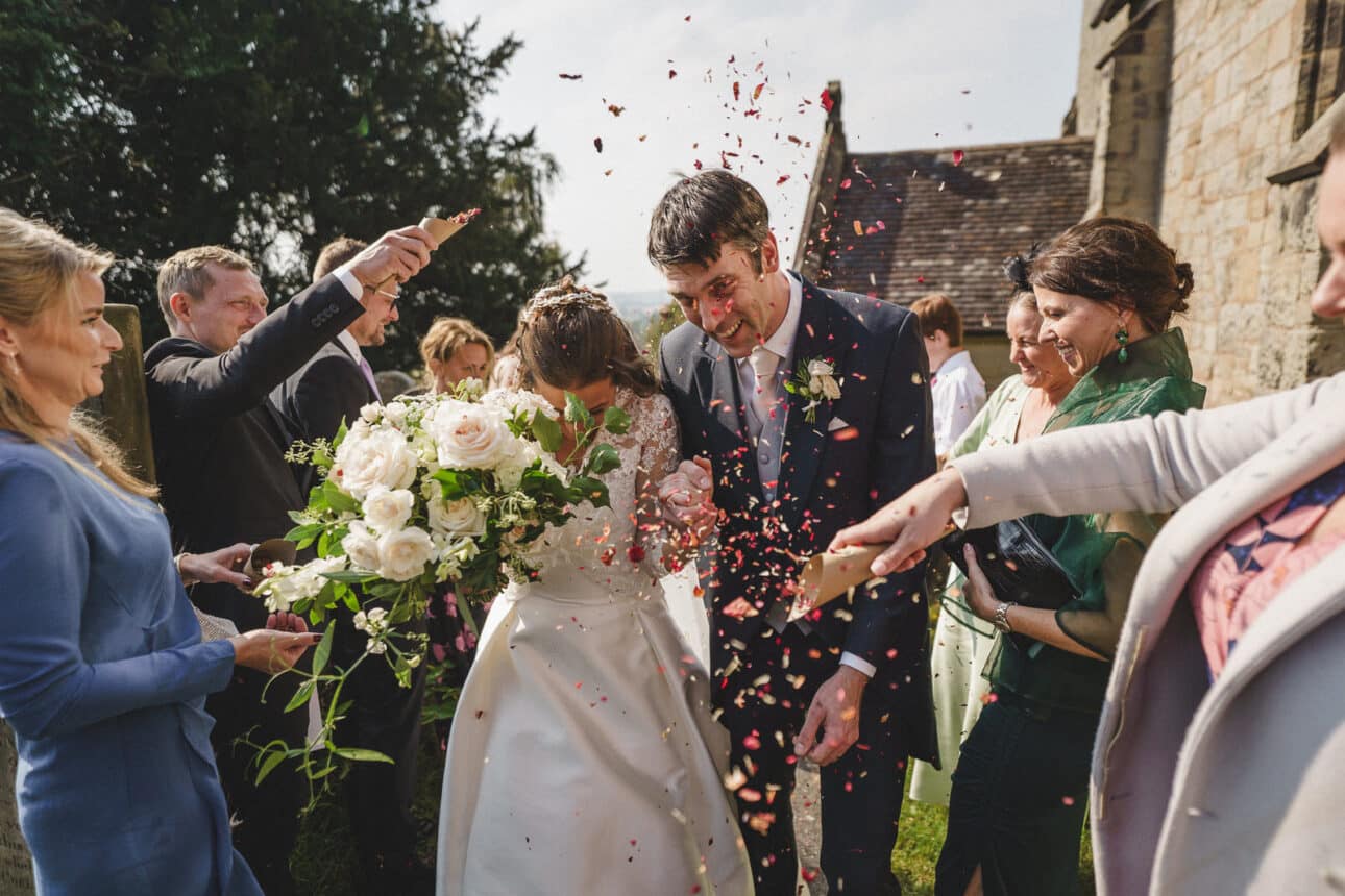 a bride and groom exit sandon hall while guests throw confetti at them; the bride holds a large bouquet of white flowers, capturing the joy of their wedding day.
