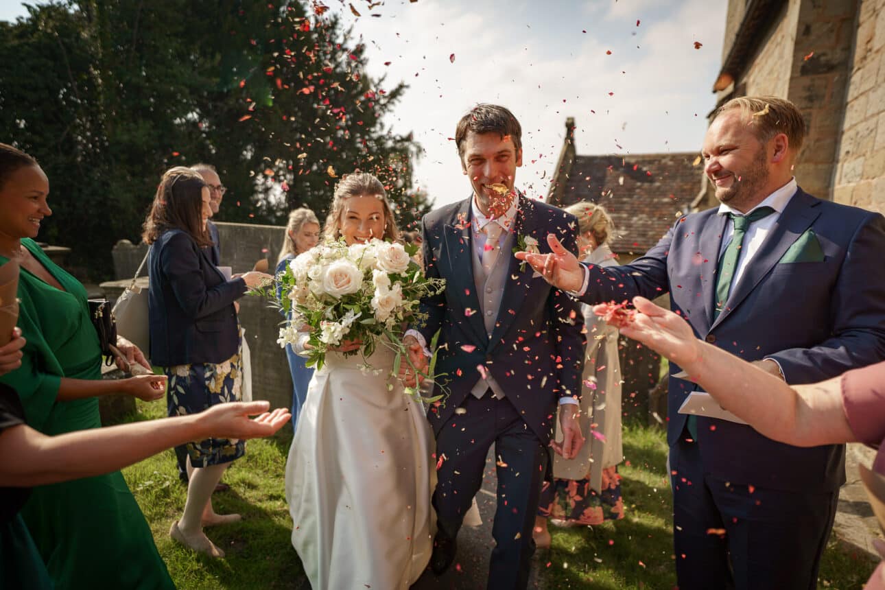 the bride and groom walk outside sandon hall as guests throw confetti at them, smiling and holding a bouquet, after their beautiful wedding day ceremony.