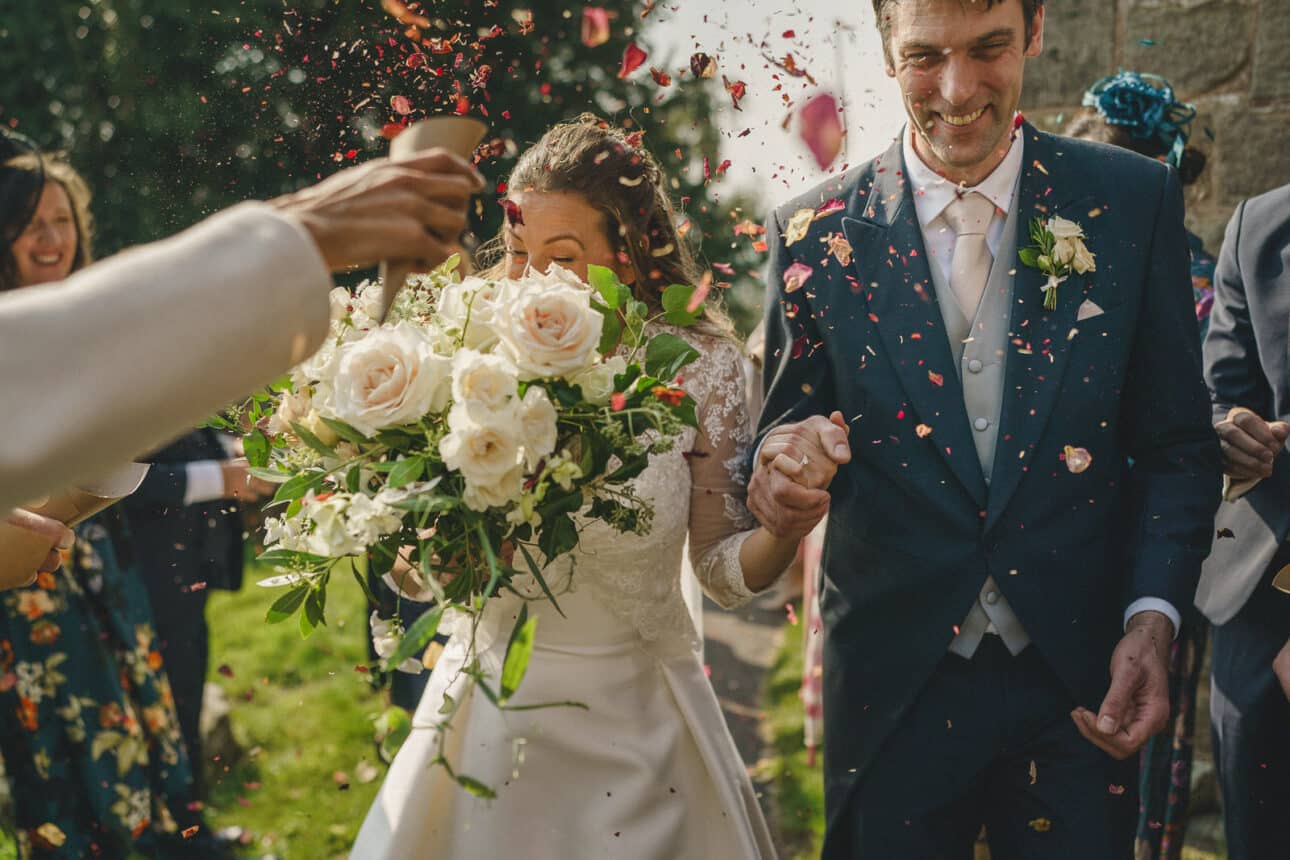 a newlywed couple holding hands walks outside sandon hall as guests toss confetti; the bride holds a large bouquet of white flowers.