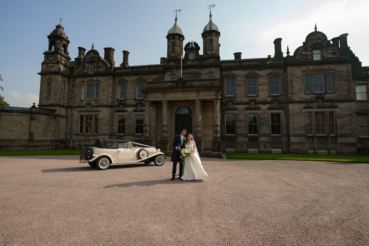 a bride and groom stand in front of a vintage car outside the grand sandon hall, a large historic stone building with ornate towers and columns, capturing their magical wedding day.
