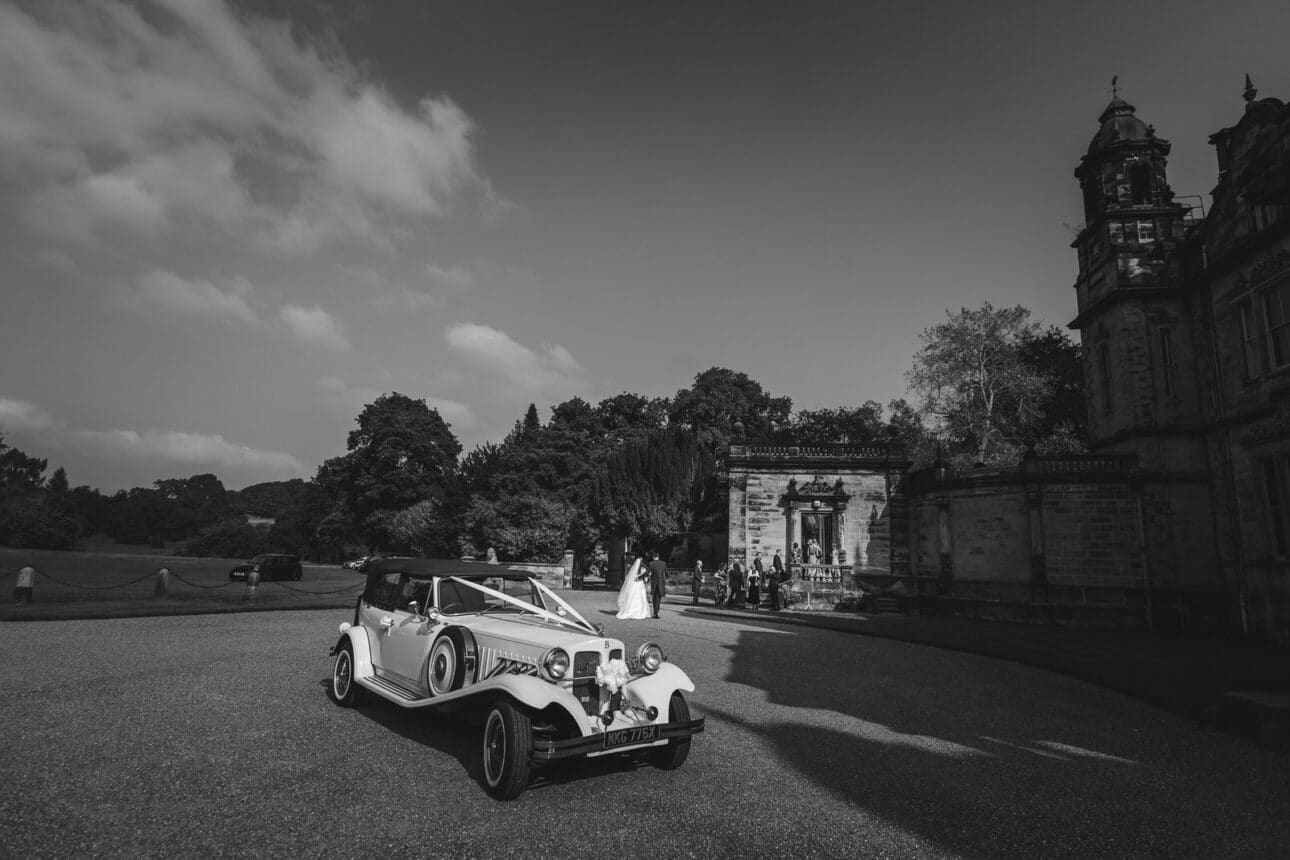 a vintage wedding car decorated with ribbons is parked outside the grand sandon hall, with people gathered in the background.