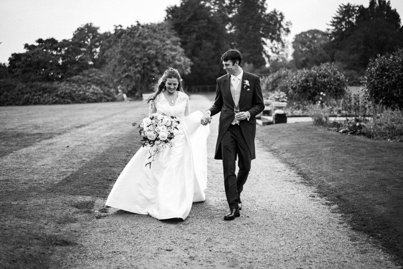 a bride and groom walk outdoors on a gravel path at sandon hall, holding hands and smiling, surrounded by greenery on their wedding day.