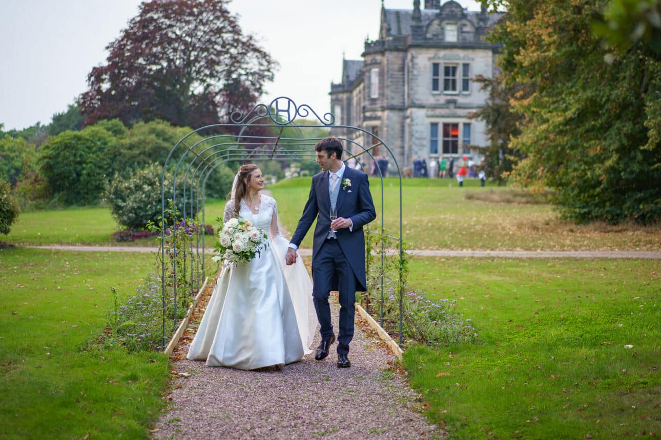 a bride and groom walk hand in hand through a garden archway, both smiling, with the grand sandon hall as a stunning backdrop—a perfect moment from their sandon hall wedding.
