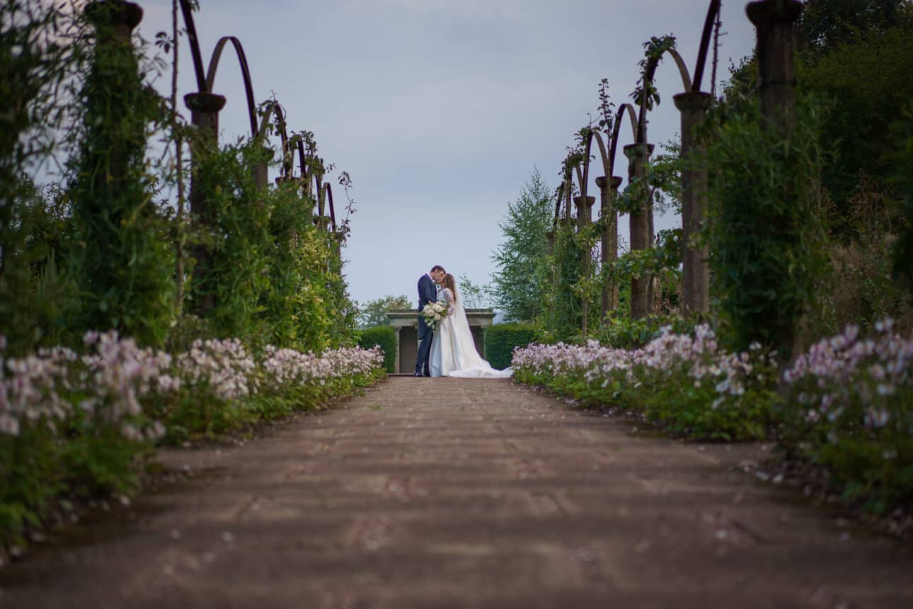 a bride and groom stand together at the end of a flower lined garden path at sandon hall, surrounded by lush greenery and arched trellises.
