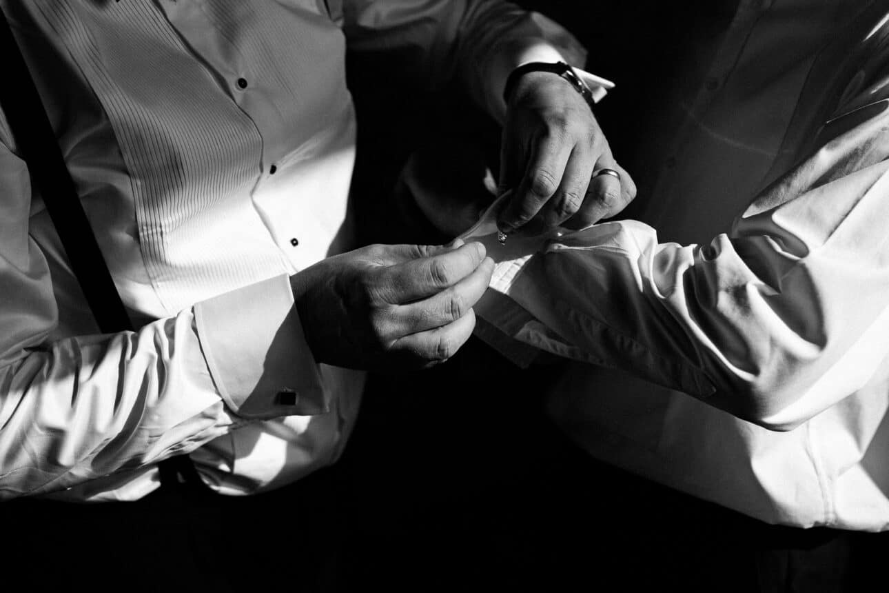 two people in dress shirts, one helping the other fasten a cufflink on a shirt sleeve, with dramatic lighting highlighting their hands—a refined moment before a sandon hall wedding.