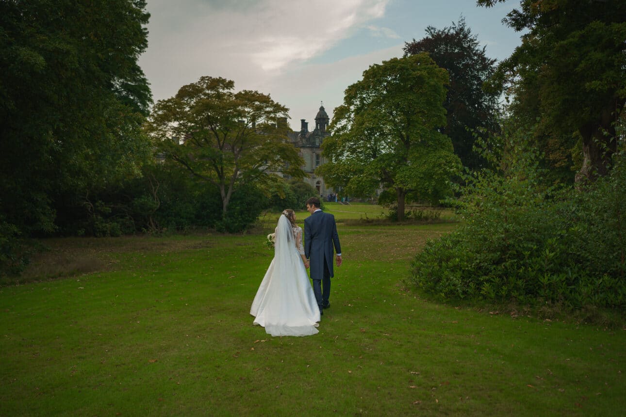 a bride and groom walk hand in hand across a grassy lawn toward sandon hall, a large, historic building surrounded by trees.