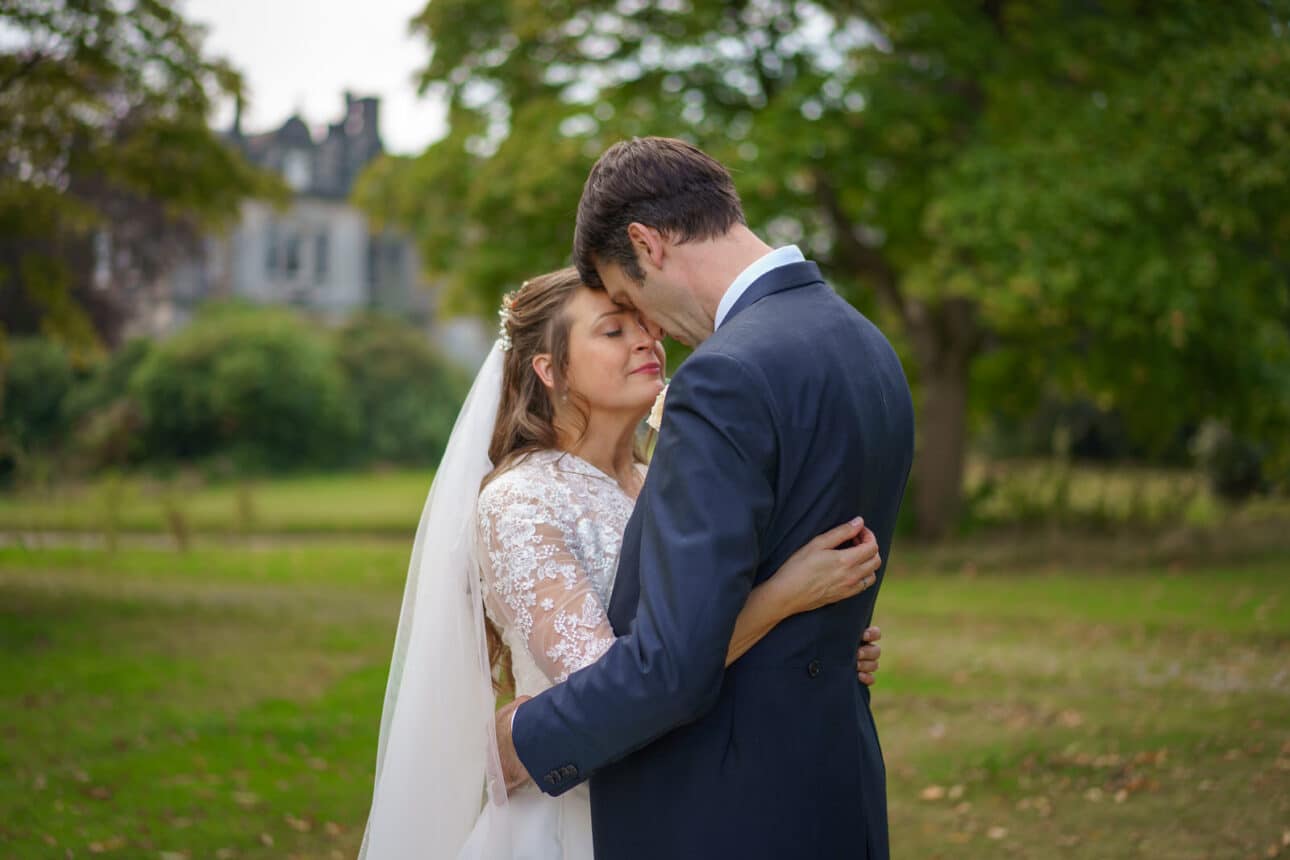 a bride and groom embrace outdoors in the gardens of sandon hall, with the bride in a white dress and veil and the groom in a dark suit, surrounded by lush greenery and towering trees.