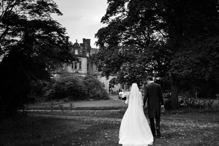 a bride and groom walk outside towards the grand stone sandon hall, surrounded by trees, captured in timeless black and white on their wedding day.