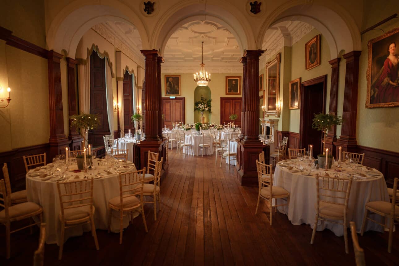 elegant dining room set for an event at sandon hall, with round tables, white tablecloths, candles, chandeliers, a wooden floor, and framed portraits on the walls—perfect for a memorable sandon hall wedding.