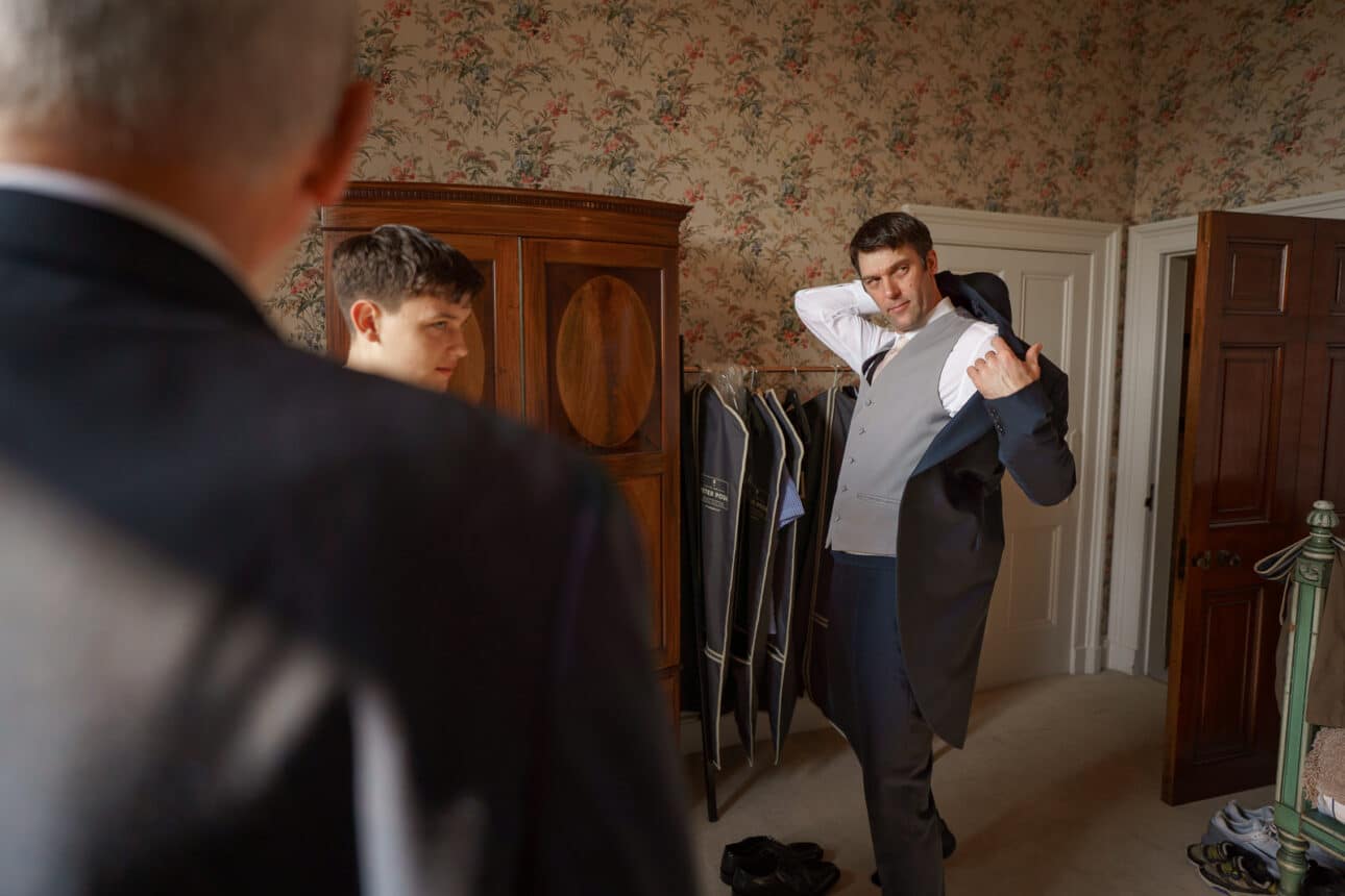 three men in suits are in a room at sandon hall; one man is putting on a formal jacket in front of a wardrobe while the others watch him, preparing for the sandon hall wedding.