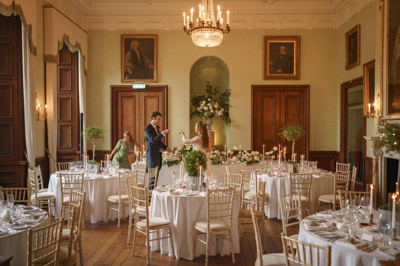 a couple stands in the center of an elegant, chandelier lit sandon hall dining room with round tables set for a formal event; two other guests are present in the background, creating a classic sandon hall wedding atmosphere.