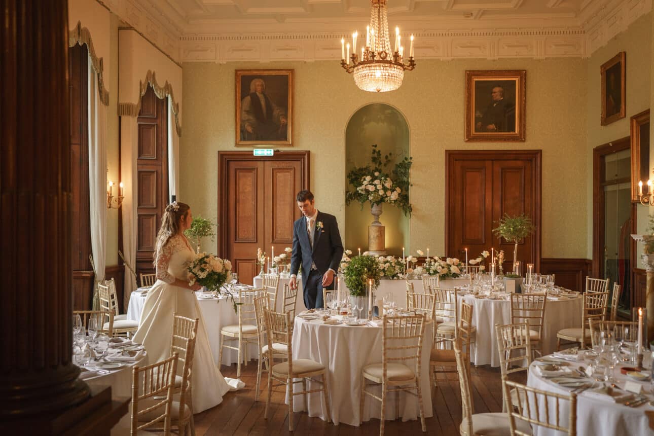 a bride and groom stand among decorated round tables in an elegant, chandelier lit dining room with wood paneling and portraits on the walls, capturing the timeless charm of a sandon hall wedding.