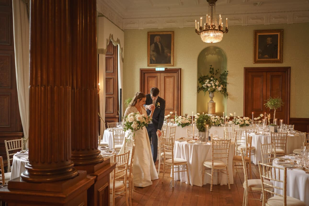 bride and groom stand together in an elegant, formal dining room at sandon hall, with white clothed tables, floral centerpieces, and classical portraits on the walls, capturing the timeless beauty of a sandon hall wedding.