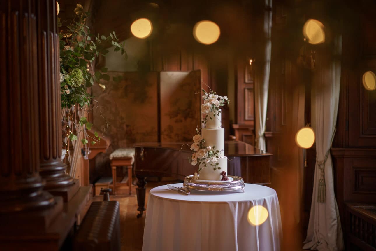 a three tiered white wedding cake with floral decorations sits on a round table covered with a white tablecloth in an ornate, wood paneled room at sandon hall, bathed in soft, warm lighting—the perfect scene for a sandon hall wedding.