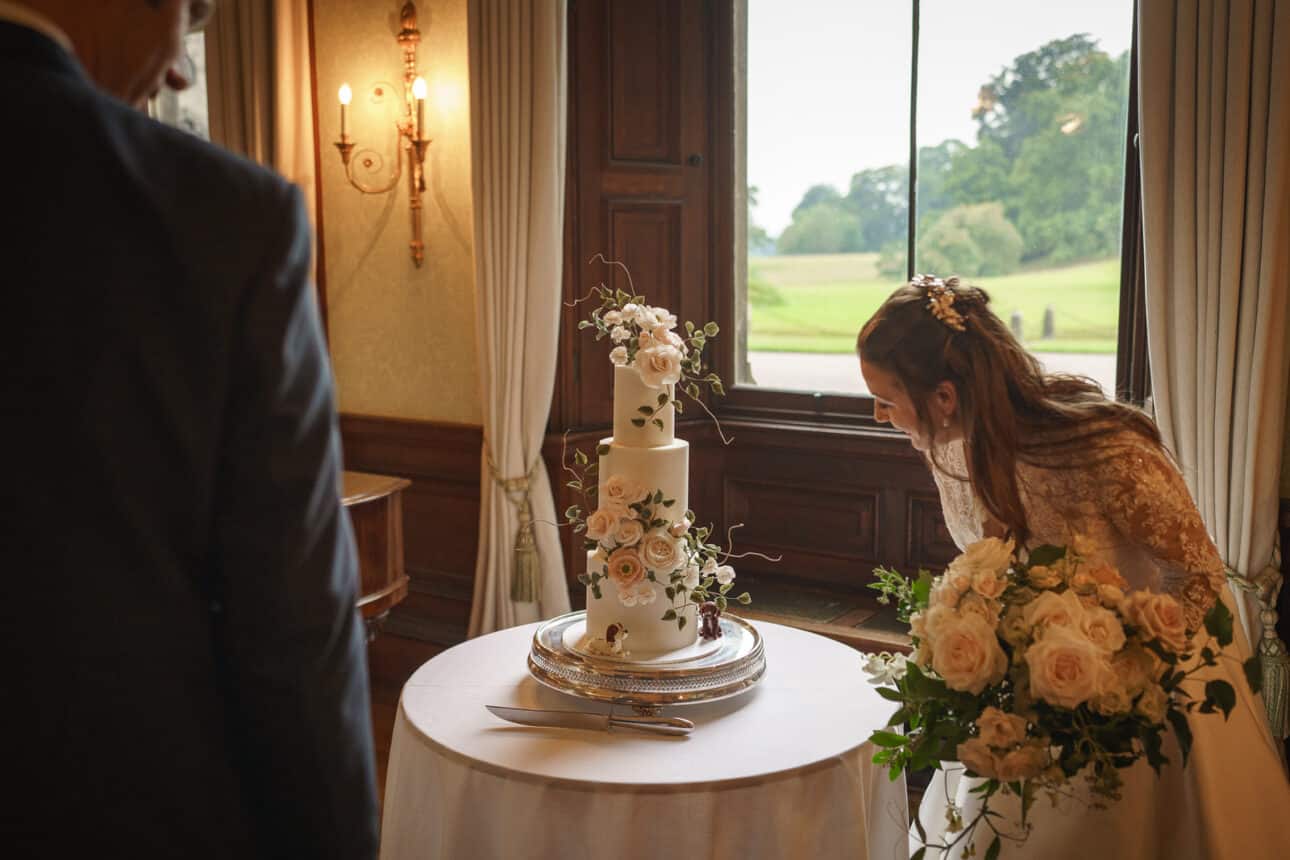 a bride holding a bouquet looks closely at a three tier wedding cake decorated with flowers at a sandon hall wedding, while another person stands nearby in the elegant, formal setting.